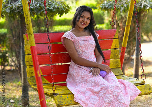 Indian young woman black long hair in pink dress wearing, she is sitting for take a photoshoot on swing is happy. isolated on beautiful park background.