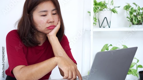 Asian woman looking bored, thoughtful, or unmotivated while working on a laptop at a bright home office desk, representing procrastination