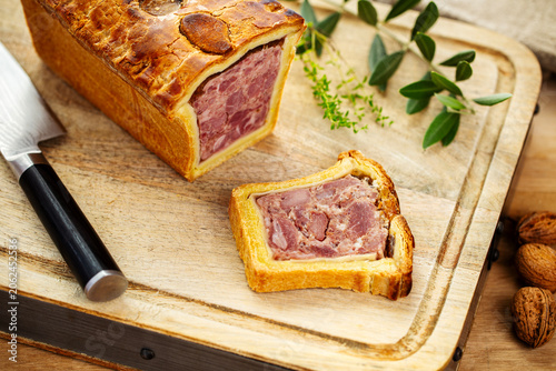 Pate en croute or pâté en croûte, with Thyme and olive tree twigs and nuts on a vintage wooden cutting board aside a black knife on wooden table.