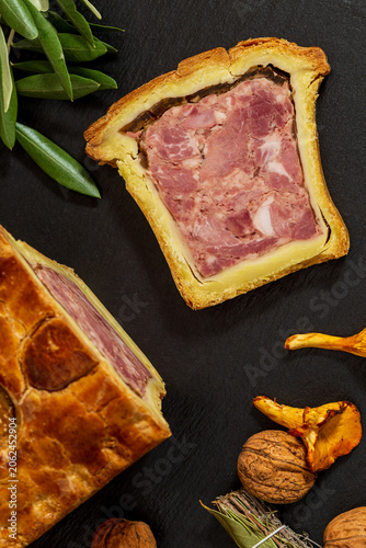 Top view of a Pate en croute or pâté en croûte and a slice of it, with chanterelles mushrooms, olive tree twig, bouquet garni and nuts on slate. Countryside vintage atmosphere.