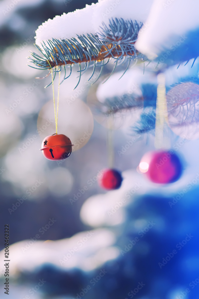 Fototapeta premium Red Christmas bell hangs on a snow-covered branch of a Christmas tree against a festive background of white snow. Christmas tree with decoration under snow
