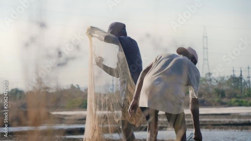 Daylight outdoor shot of two Indian Asian male fishermen untangling, preparing, and organising a fishing net before casting it into a lake or pond water in search of a catch on a sunny but windy day. 