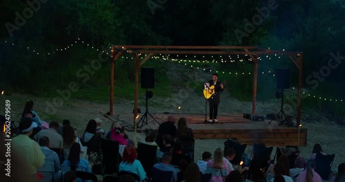 Solo acoustic guitarist performing on a string-lit pergola stage at dusk