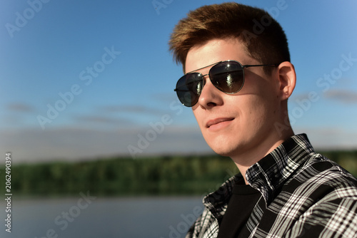 A handsome young man with dark hair and sunglasses poses confidently by the lake on a sunny day.