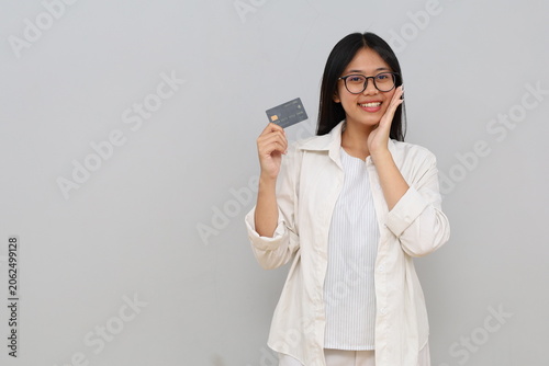 Photo of an impressed young Asian woman, hand on cheek, holding credit card, smiling looking at camera, isolated on white background.