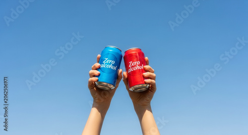 Hands holding up a Zero alcohol drink can in blue and red variations, symbolizing healthy refreshment and sober choices under a blue sky.