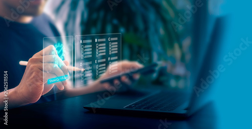 A person uses a stylus to touch a virtual screen while holding a smartphone in one hand digital checklist and quality badge icon. A laptop sits on the desk in a bright room filled with plants.