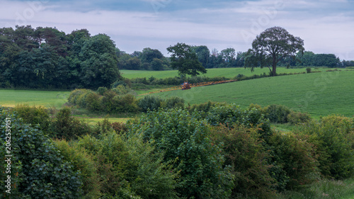 Lush green fields stretch beneath a soft, cloudy sky. Lone tractor and sprayer in the far distance through the meadow. Trees frame the landscape, offering quiet shelter. Nature's calmness invites peac