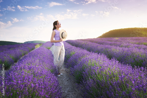 Young woman enjoy lavender field and holding a straw hat at the sunset.