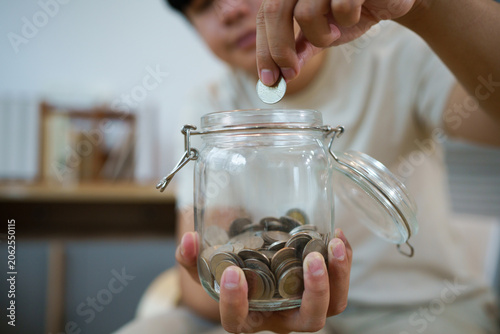 Young Asian Person Dropping Coin into Glass Jar Full of Coins for Savings at Home