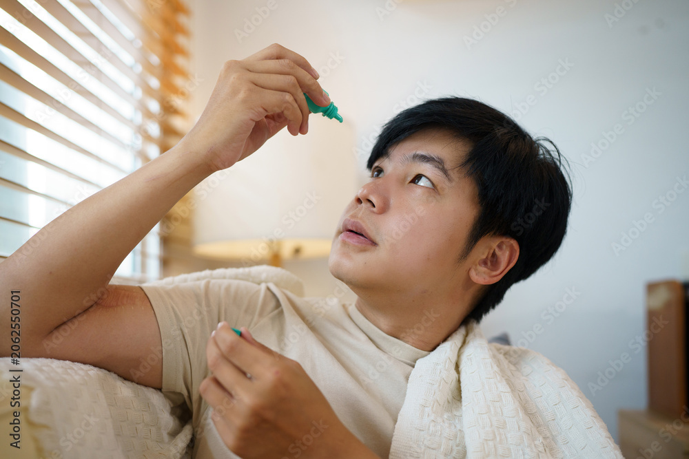Naklejka premium Young Asian Man Applying Eye Drops at Home for Dry Eye Relief or Allergy Treatment