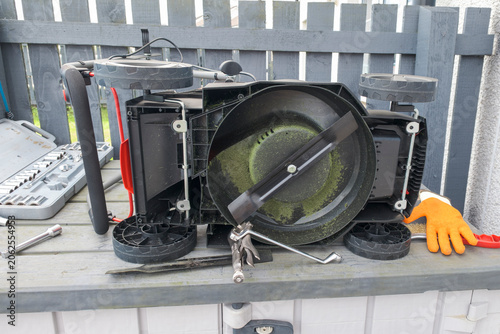 Broken Lawn mower rests on a workbench outdoors. Tools and gloves nearby suggest repair or disassembly. Fence behind adds urban or backyard setting. Image conveys technical, hands-on