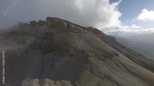 Dynamic FPV drone shot racing along a rocky high mountain ridge with mist rolling over the crest and sunlight breaking through, capturing wild alpine terrain and a steep remote valley below