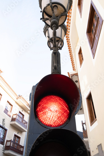 bright red traffic light glows, commanding a halt. The low-angle view captures the signal's intense light against a clear sky, with an ornate lamppost and a building behind.