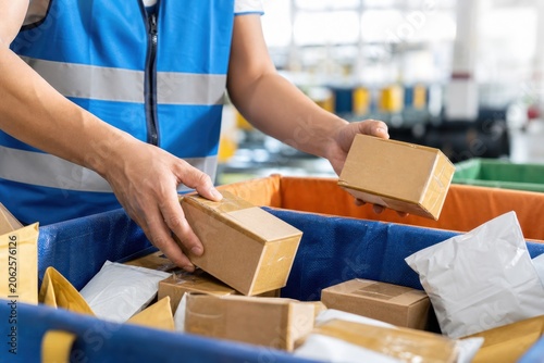 Professional warehouse worker sorting cardboard boxes and mail packages into bins inside distribution center, close-up view of hands, logistics concept