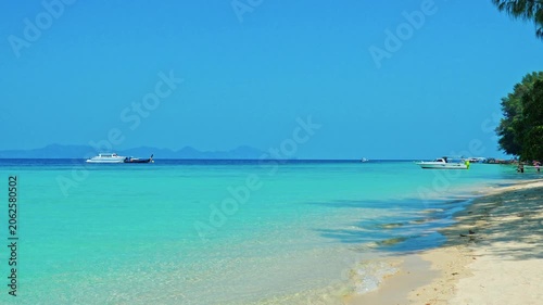 Shoreline meeting turquoise ocean water under clear blue sky, white sand beach with parked boats on horizon, tropical island tourism and travel advertising background