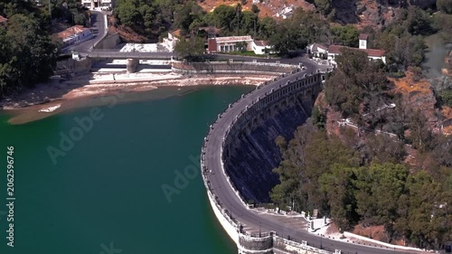Drone footage advancing over El Chorro's elegant curved dam structure, tracing its arched stone parapet beside deep emerald water, wooded banks, and a whitewashed keeper's house above.