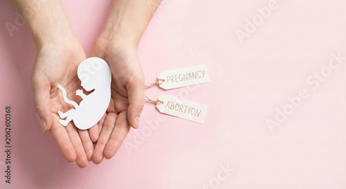 Female hands holding paper fetus silhouette on pink background with cards symbolizing reproductive rights and difficult life choice decision
