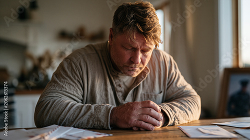 Middle-aged male veteran sits alone at a kitchen table at dusk, head bowed and hands folded, an expression of quiet moral weight crossing his face as unopened mail and a framed