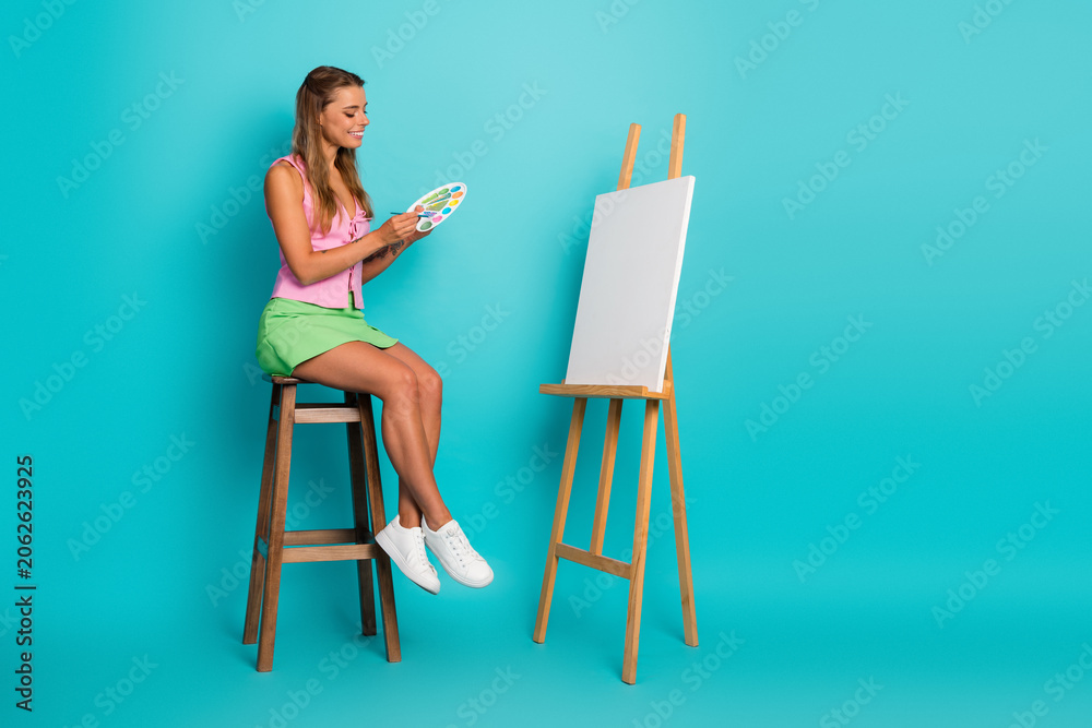 Fototapeta premium Young woman painting on a canvas in a bright blue studio background with a smile and summer chic vibe