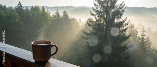 Close-up of a cup with water vapor on a wooden railing against a foggy forest and sunbeams.