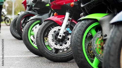 Close up view of several parked motorcycles lined up, featuring vibrant green and red fairings, highlighting suspension, tire treads, and disc brakes on a wet ground surface outdoors.