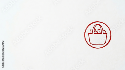 Red ink stamp of a shopping basket icon on a white textured background. Minimalist abstract symbol of a bag filled with round objects. Copy space for text