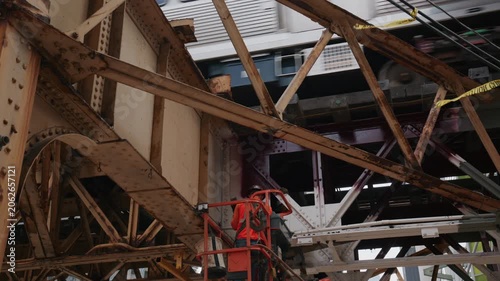 Construction worker in boom lift grinding rusty steel girder of elevated Chicago L train track while public transit passes on bridge structure in city