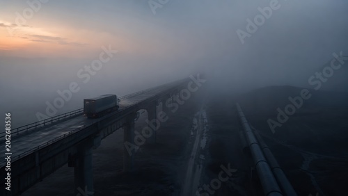 A lone vehicle traverses a misty viaduct at dawn, shrouded in atmospheric fog.