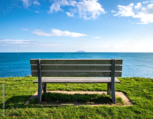 Wooden park bench on green grass overlooking a blue ocean with an island on a clear, sunny day
