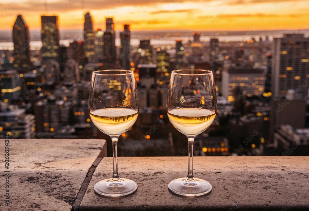 Obraz premium Two glasses of white wine on a stone ledge overlooking a city skyline at sunset. Golden hour view of urban skyscrapers from a rooftop. Romantic evening and travel concept