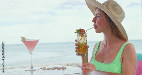 Young woman drinking passion fruit Passiflora edulis cocktail through straw, luxury beach resort overlooking ocean, tropical summer vacation setting, travel leisure lifestyle