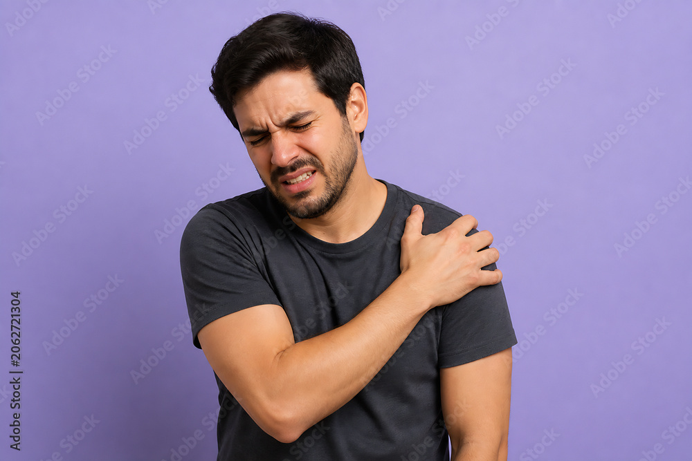 Fototapeta premium A man is clutching his shoulder in apparent pain against a plain purple background.