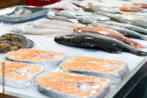 A traditional Taiwanese fish market stall showcases fresh seafood displayed on crushed ice, including whole snappers, dark-scaled fish, and vibrant salmon slices. The composition highlights freshness,