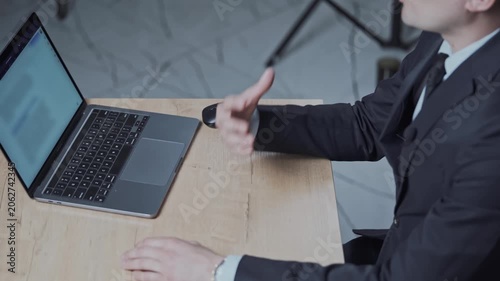 Man speaks by gesturing with his hands. Person in a black suit and tie is sitting at a table with a laptop. Businessman or an office worker advises clients. Conclusion of the transaction.