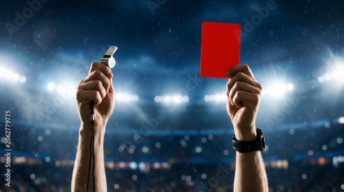 Referee holding a red card and whistle high in a professional soccer stadium during a night match under bright floodlights to indicate a penalty or player expulsion during competitive play
