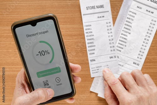Smartphone showing discount next to receipts, hands holding printed store receipts while loyalty app displays badge, shopper comparing totals and scanning coupons, focus