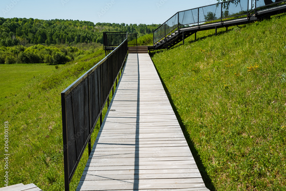 Fototapeta premium Serene outdoor walkway. Tranquil pathway with scenic views. Gentle hill trail shaded by calming summer sunlight. Calm journey along wooden route bordered by dark railing during summer