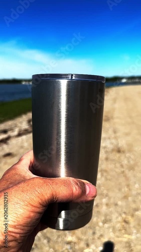 Close-up of a man's hand holding a reusable stainless steel travel mug on a sunny day. The background shows a gravel path near water