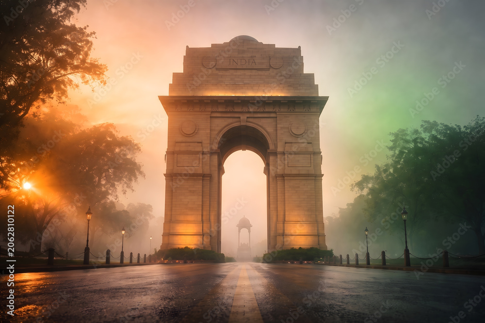 Naklejka premium Majestic India Gate New Delhi Surrounded by Dramatic Tricolor Saffron White Green Misty Fog at Dawn with Wet Road Reflection and Glowing Street Lamps — India Independence Day Republic Day Patriotic