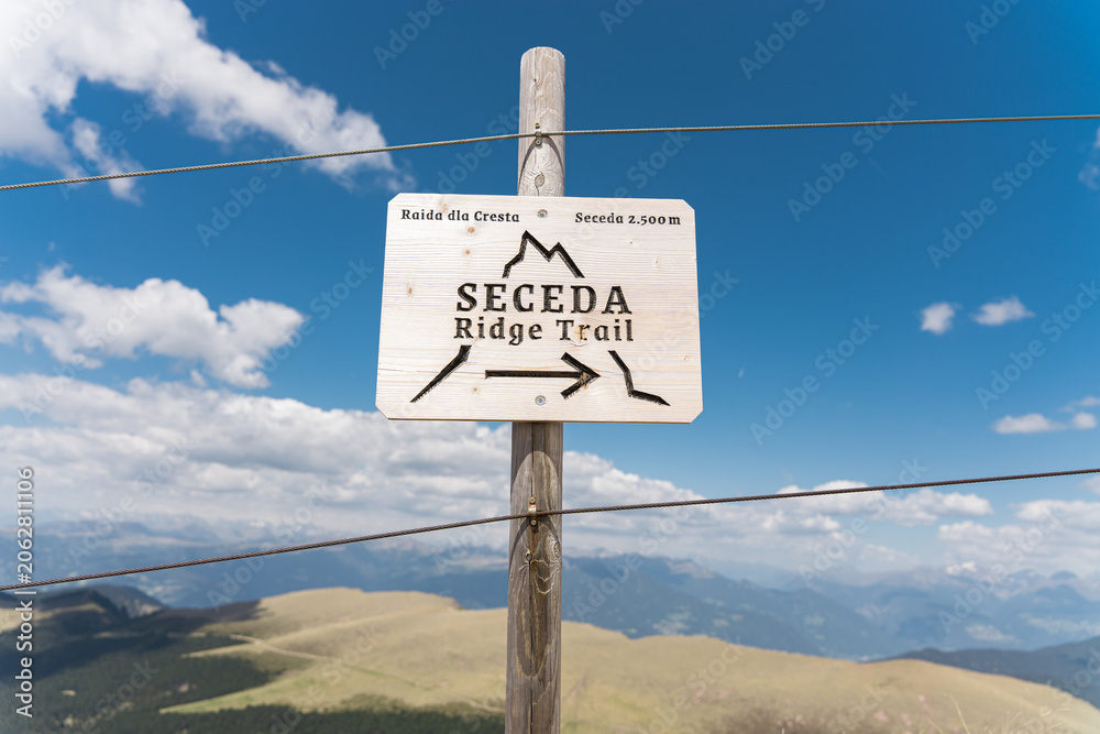 Naklejka premium Wooden directional sign for Seceda Ridge Trail on a mountain pass with panoramic alpine landscape view under blue sky, outdoor hiking and trekking navigation concept