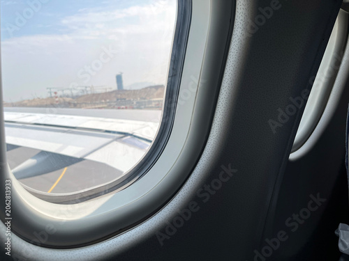 View from an airplane window showing the aircraft wing, runway, airport terminal, and distant control tower under a bright sky. Travel and aviation scene captured from inside the cabin before takeoff 
