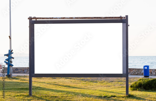 Large horizontal blank white outdoor advertising billboard mockup on a green grass field near a coastal promenade with sea view under a bright evening sky.