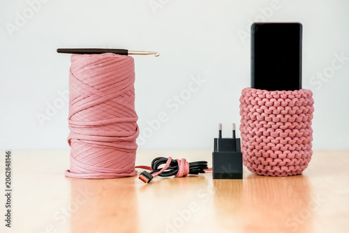 Smartphone in a pink knitted pouch next to a yarn ball and charger. Concept of digital detox, offline lifestyle, creative handmade hobbies, relaxation and slow living.