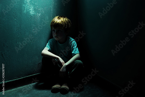 Child sitting alone in a dark room, looking towards an open door with light, conveys themes of fear, hope, and vulnerability.