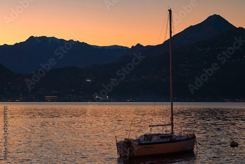 A solitary sailboat rests on tranquil waters as the sun dips below silhouetted mountains, casting a warm, orange glow across the sky and water