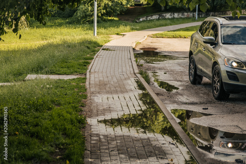 Foto Silver car parked by a wet sidewalk with puddles, grass, and leafy trees in warm sunlight
