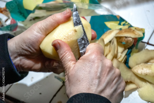 Primer plano de manos de mujer mayo pelando una patata con un cuchillo de cocina pequeño, mostrando las peladuras en el fondo durante el proceso de preparación de ingredientes caseros tradicionales.