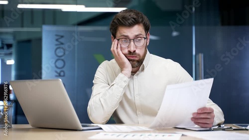 Confused businessman sitting at desk in office struggling with paperwork and looking at laptop screen. Frustrated company owner reviewing documents, facing accounting mistakes or business problems.