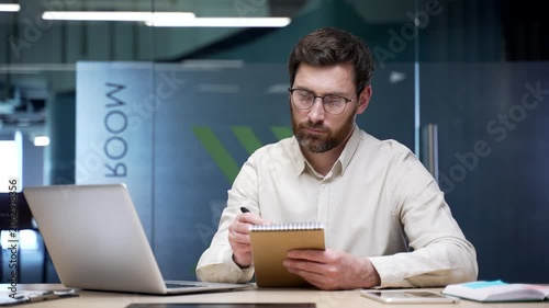 Businessman watching video conference on laptop and taking notes in notebook sitting in office. Focused professional attending virtual training, e-learning course, online seminar for skill development